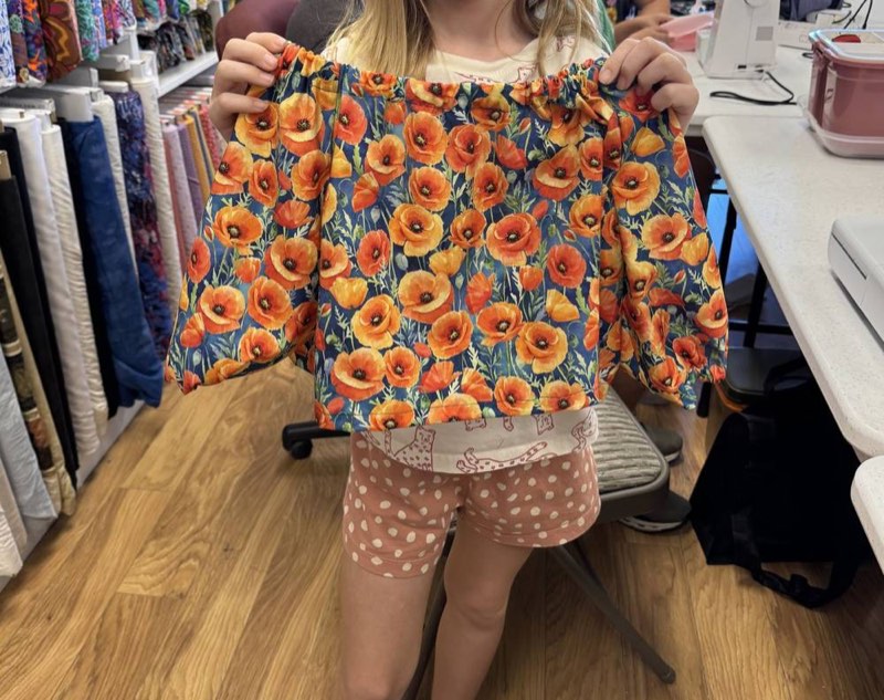 A young student holding up a finished orange poppy-print top she sewed.